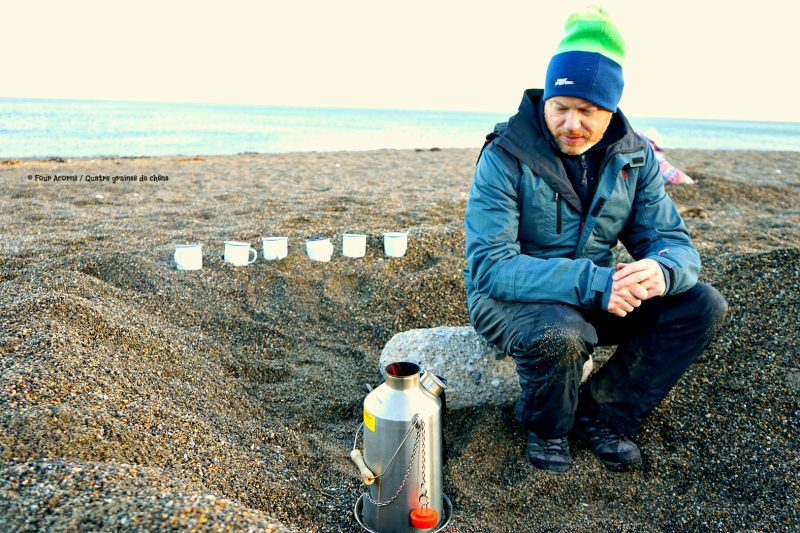 man-sitting-rock-kelly-kettle-six-white-cups-sand-beach